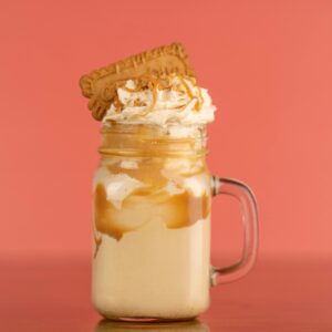 Close-up of a caramel milkshake with whipped cream and biscuit topping against a salmon background.