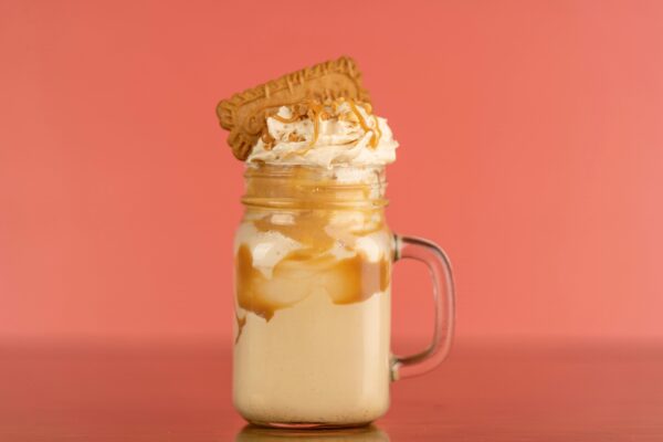 Close-up of a caramel milkshake with whipped cream and biscuit topping against a salmon background.
